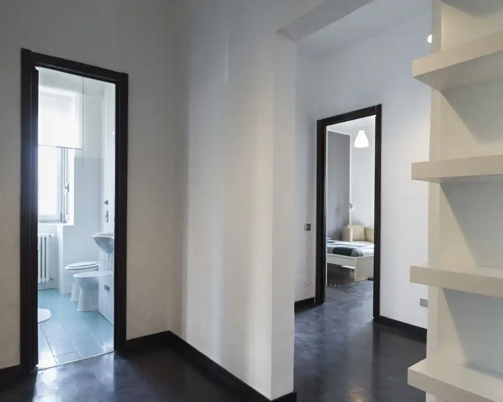 Ingresso/corridoio luminoso e minimalista con pavimento scuro, pareti bianche e scaffalatura integrata che conducono al bagno e alla camera da letto.