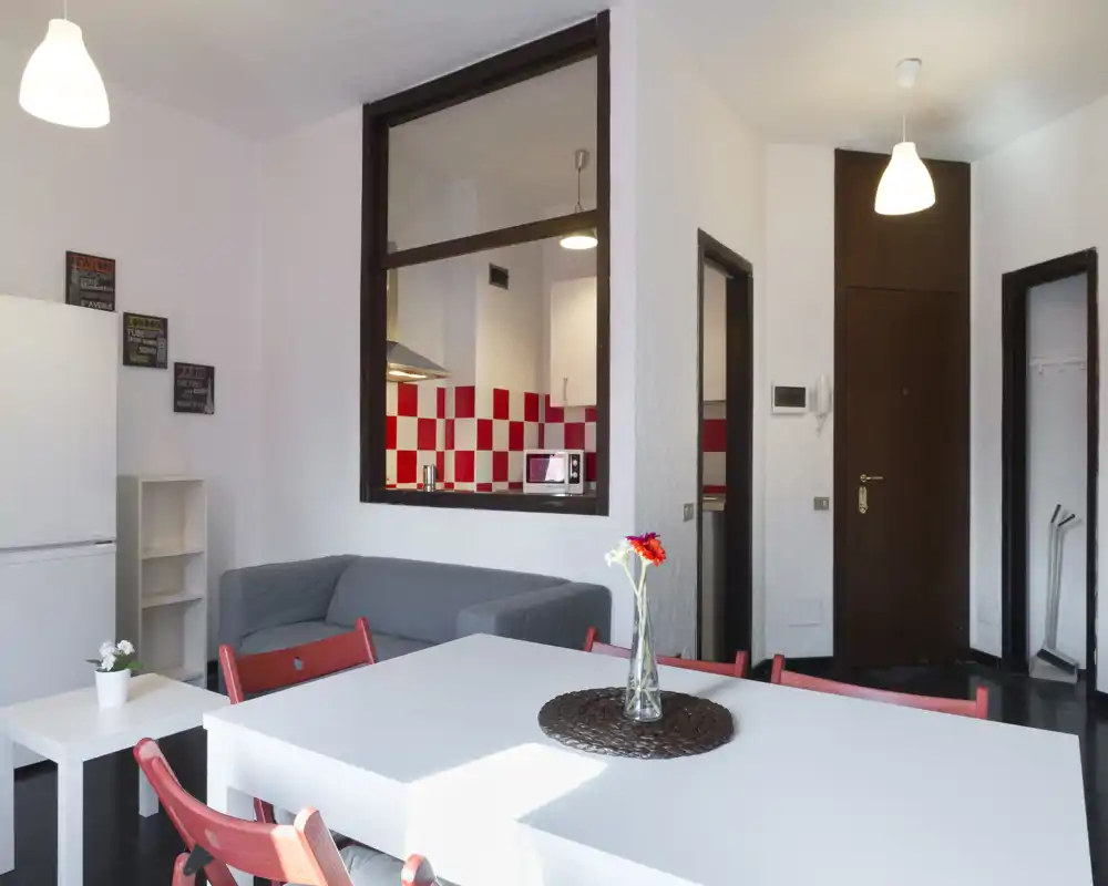 Zona giorno luminosa e accogliente con tavolo da pranzo e divano grigio; una finestra passante mostra la cucina compatta con piastrelle rosse e bianche.