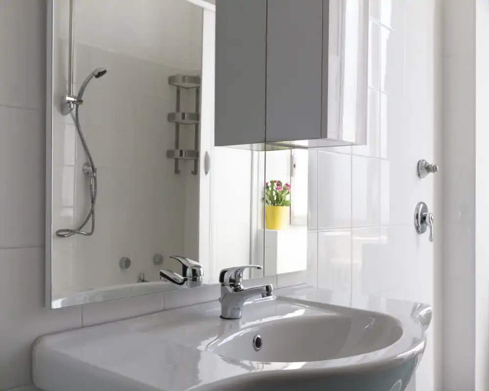 Lavabo moderno e luminoso con miscelatore cromato, specchio e accessori della doccia visibili; piastrelle bianche e stile minimal pulito.