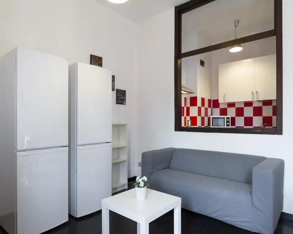 Area giorno semplice e moderna con divano grigio, piccolo tavolino bianco e vista diretta sulla cucina con piastrelle rosse e bianche. Spazio luminoso, minimal e funzionale adatto a soggiorni brevi.