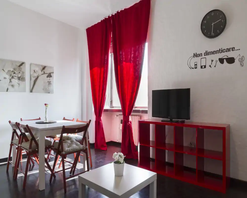 Zona giorno luminosa e moderna con tende rosse intense e arredi coordinati. L'ambiente comprende un tavolo da pranzo, una TV su una scaffalatura rossa e dettagli decorativi semplici per un'atmosfera accogliente.