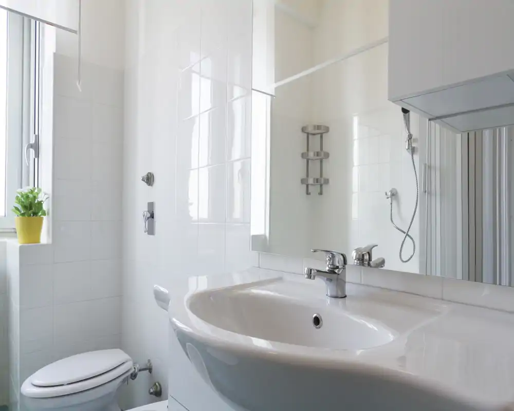Bagno moderno e luminoso con lavabo ampio, specchio a parete e doccia visibile nello specchio. Piastrelle bianche e luce naturale donano un aspetto pulito e arioso.