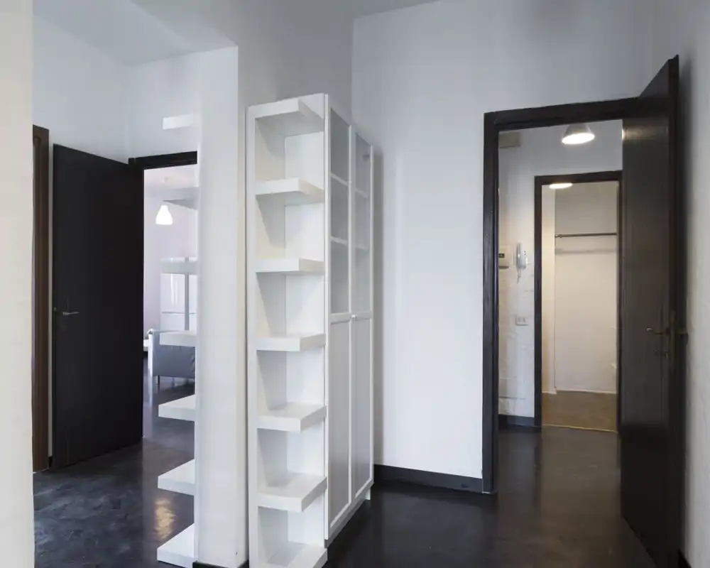 Ingresso/corridoio minimale e luminoso con pavimento scuro, pareti bianche e porte aperte che conducono ad altre stanze. Un esile mobile a giorno bianco definisce l'area.