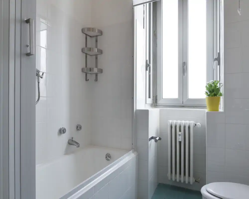 Bagno luminoso e pulito con vasca, pareti piastrellate bianche, termosifone sotto la finestra e una pianta sul davanzale. Spazio semplice e ben curato, adatto alle foto dell'annuncio.