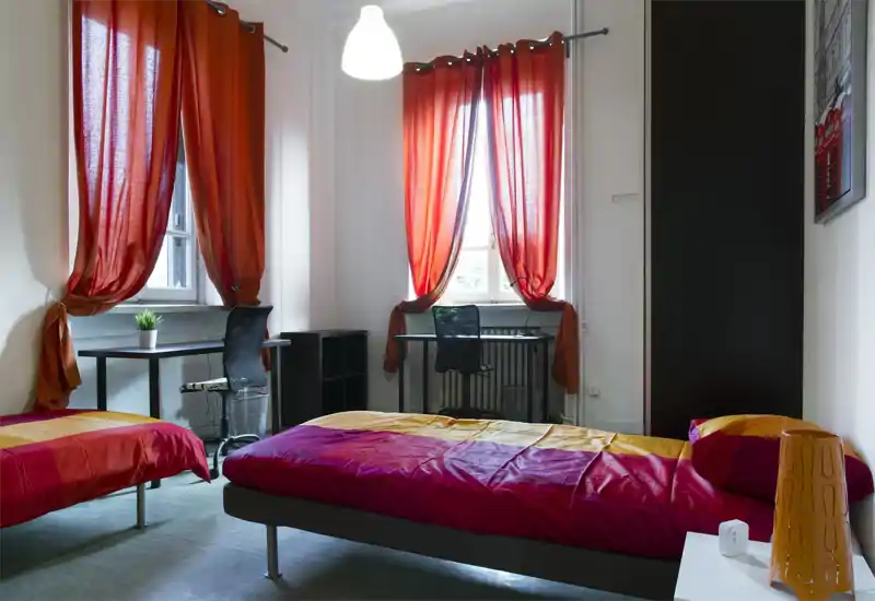 Camera con due letti singoli, grandi finestre luminose e due scrivanie. Tende rosso-arancioni e biancheria coordinata creano un ambiente accogliente e ben illuminato, ideale per studenti o affitti condivisi.