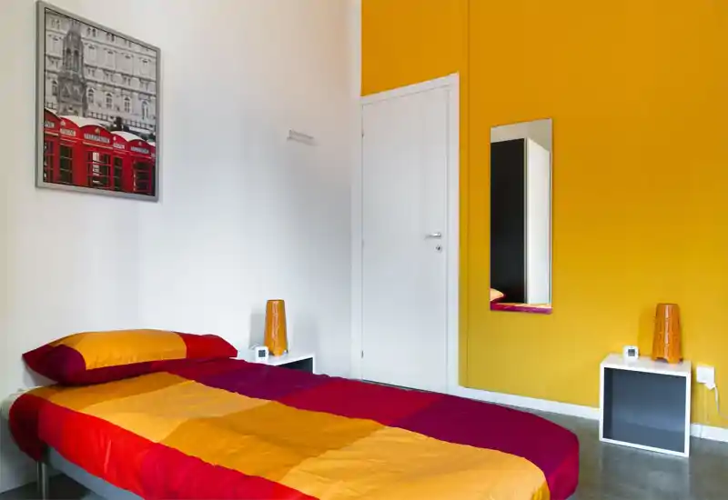 Camera da letto luminosa e moderna con copriletto arancione e rosso, due comodini con lampade coordinate e uno specchio a figura intera su una parete gialla vivace.