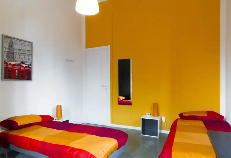 Camera da letto luminosa e moderna con due letti singoli con biancheria arancione e rossa, una parete d'accento gialla e piccoli comodini bianchi; linee pulite e colori vivaci creano un ambiente allegro e ben illuminato.