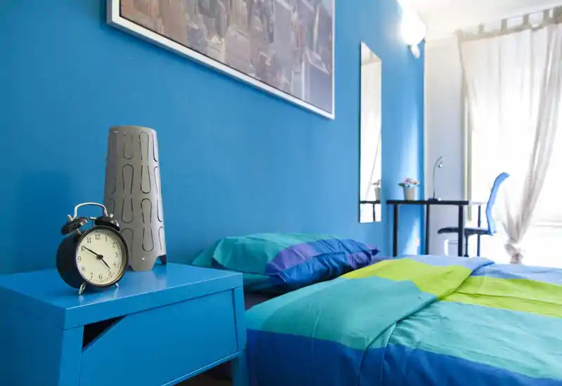 Camera da letto luminosa e colorata con letto matrimoniale ben rifatto, comodino e scrivania vicino alla finestra. Il vivace tono blu e la luce naturale creano un ambiente moderno e accogliente, perfetto per affitti brevi.
