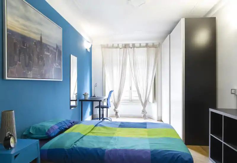 Camera da letto luminosa e colorata con letto matrimoniale, scrivania vicino alla finestra e ampio armadio. La luce naturale entra dalle tende leggere, creando un ambiente fresco e accogliente.