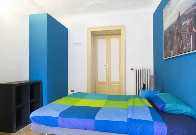 Camera da letto luminosa e moderna con copriletto blu-verde vivace, comodino essenziale e linee pulite — ideale per soggiorni singoli o in coppia.