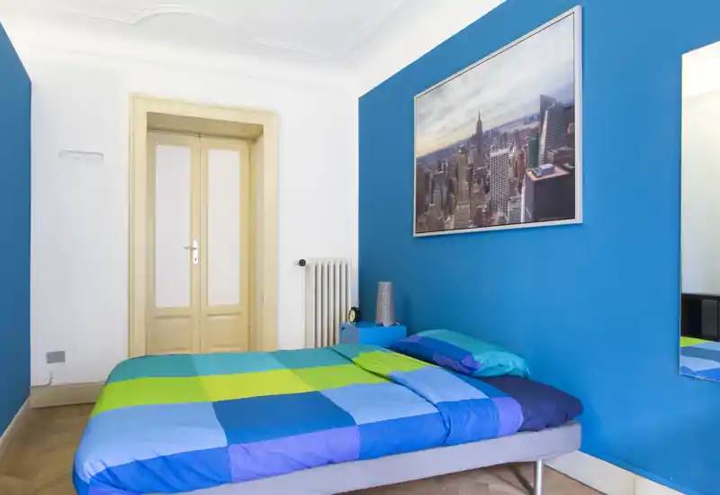 Camera da letto luminosa e moderna con letto matrimoniale coperto da biancheria colorata blu e verde, un comodino e un grande quadro di una città sulla parete blu acceso.