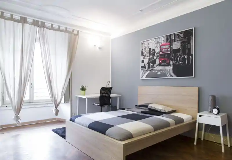 Camera da letto luminosa e moderna con letto in legno, copriletto a quadri e scrivania vicino alla finestra. Luce naturale morbida, arredamento minimal e una stampa urbana incorniciata creano un’atmosfera tranquilla e di stile, perfetta per annunci.