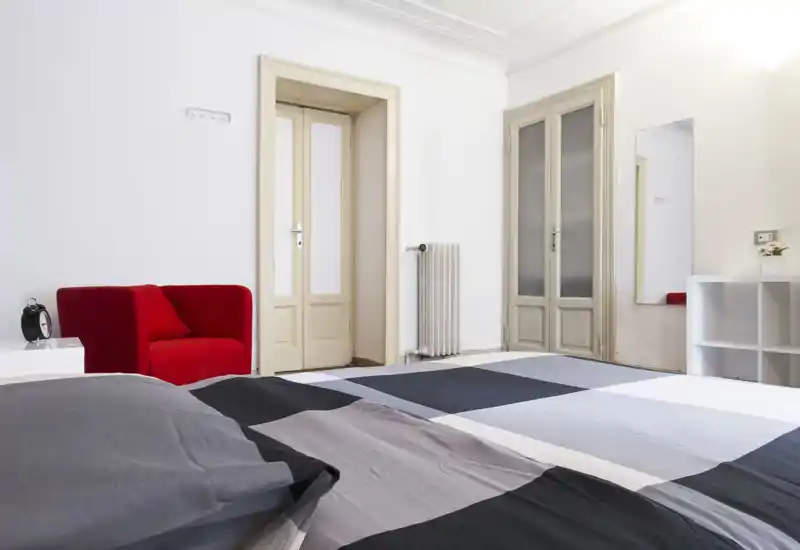 Camera da letto luminosa e minimal con un grande letto in primo piano, una poltrona rossa come elemento di accento e pareti neutre per un aspetto pulito e moderno adatto agli annunci.
