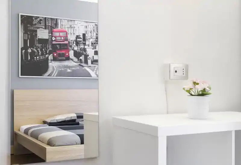 Minimalist bedroom corner with a low wooden bed seen in a wall mirror, neutral striped bedding and a framed city photo above. Bright, clean composition suitable for a listing highlighting a cozy, modern single or guest bedroom.