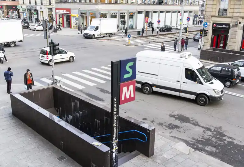Street-level outdoor view showing a subway entrance with stairs, pedestrians, vehicles and a zebra crossing in an urban setting.