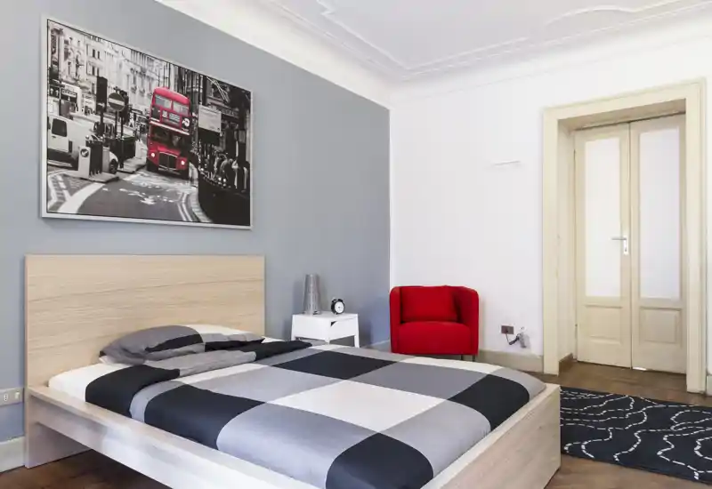 Camera da letto luminosa e moderna con letto matrimoniale, copriletto a quadri, comodino e poltrona rossa. Toni neutri e un quadro urbano donano un'atmosfera contemporanea e accogliente, ideale per affitti.