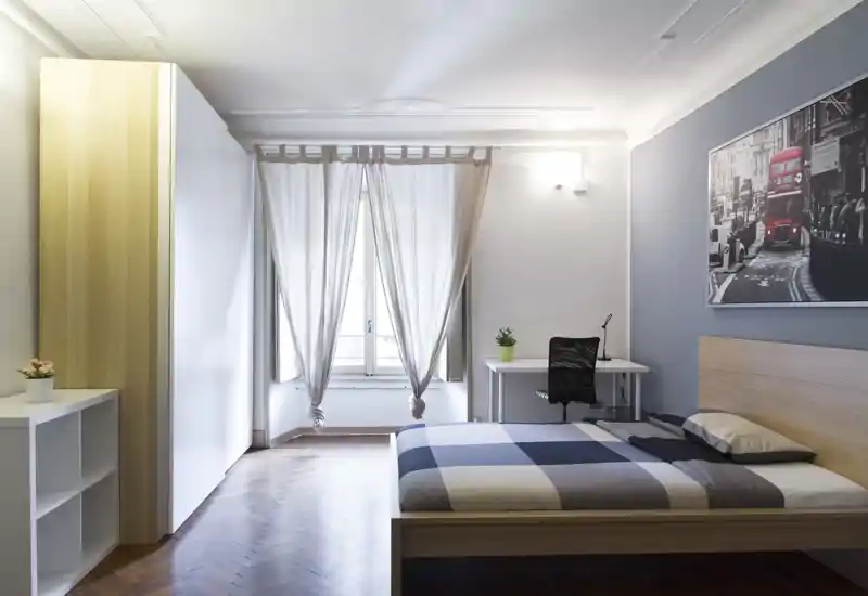 Camera da letto moderna e luminosa con letto matrimoniale, scrivania e armadio. La luce naturale entra dalla finestra con tende leggere e gli arredi minimal creano un ambiente tranquillo e accogliente.