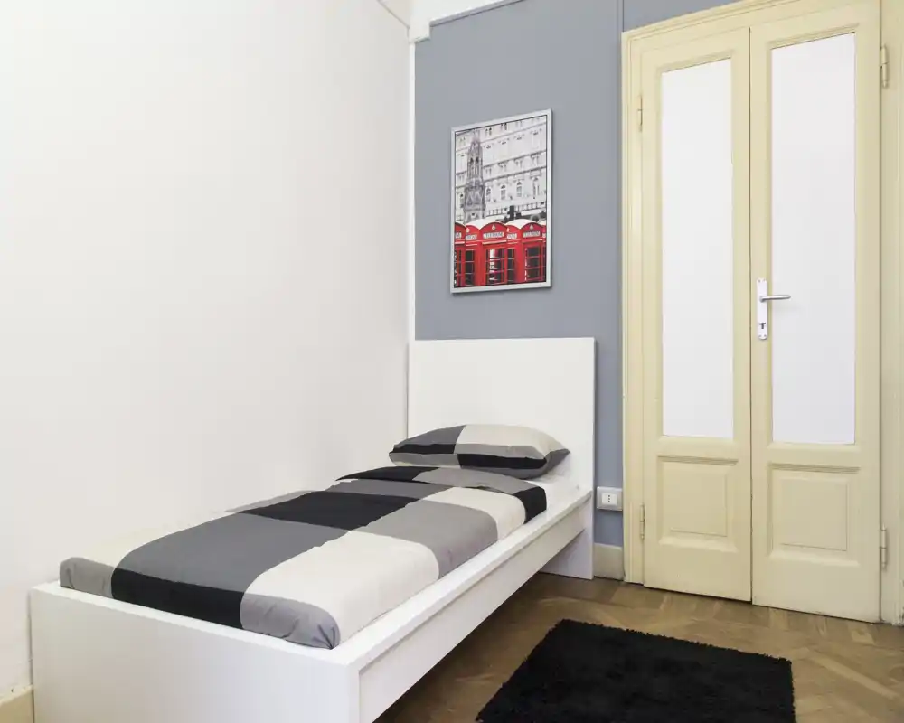Chambre simple minimaliste avec lit sur plateforme blanc, linge de lit monochrome, tableau mural et tapis noir moelleux ; lumineuse, soignée et décorée sobrement, idéale pour location courte durée.