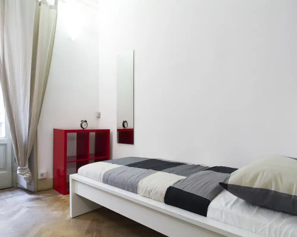 Chambre minimaliste et lumineuse avec un lit simple blanc sur plateforme, linge de lit gris et blanc et une petite étagère rouge. La lumière naturelle provenant d’une grande fenêtre donne une ambiance aérée, adaptée à une annonce de location.