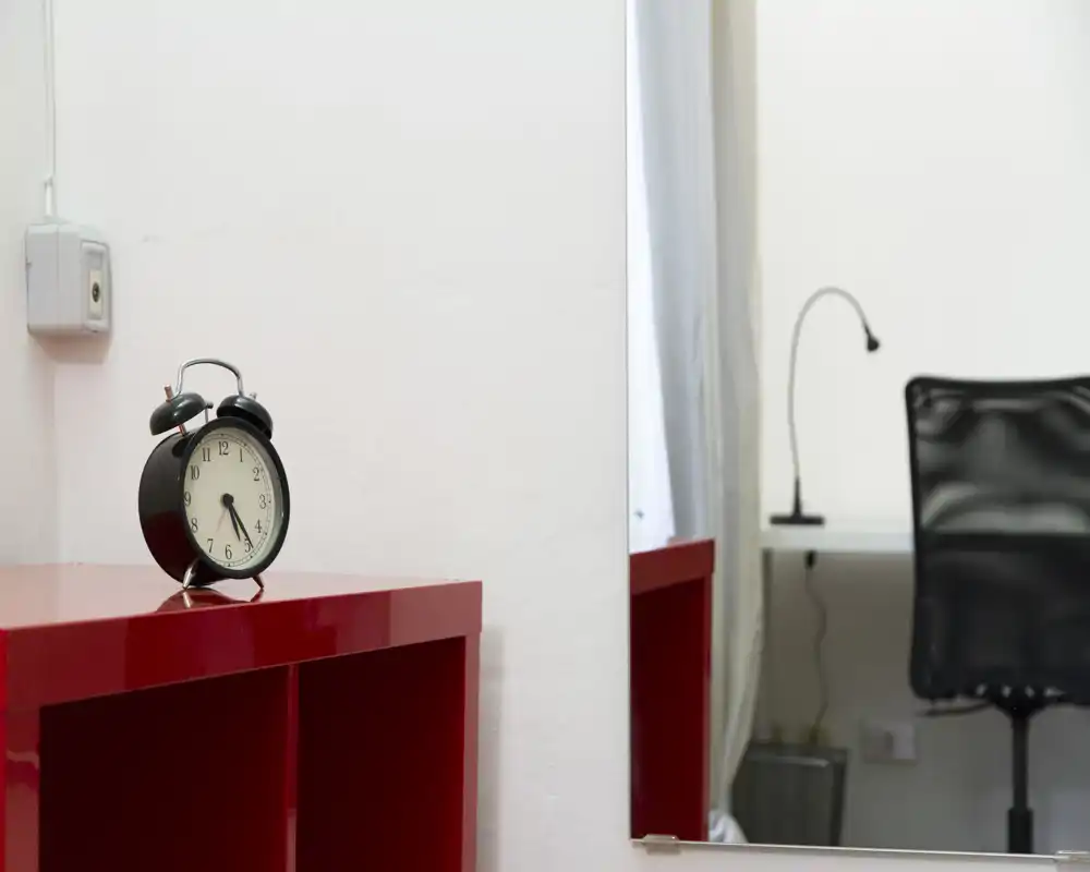 Détail d'un coin salon minimaliste avec une étagère rouge, un réveil vintage et un aperçu d'un bureau et d'une chaise reflétés dans le miroir ; coin propre et lumineux adapté à une annonce locative compacte.