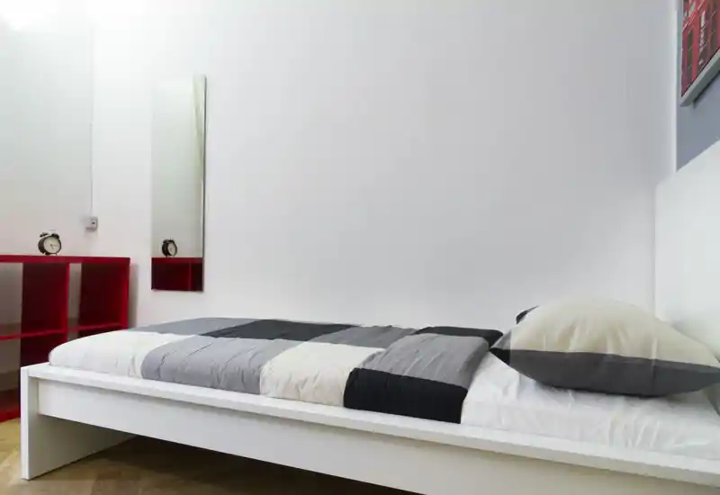 Camera da letto singola luminosa e minimal con struttura letto bianca bassa e copriletto grigio a blocchi. Arredamento moderno essenziale, specchio stretto e scaffale rosso che crea un contrasto.