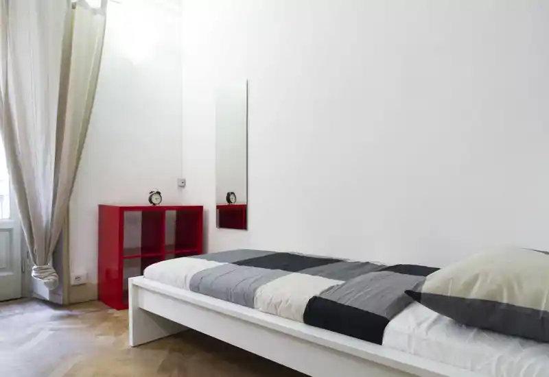 Camera da letto minimal e luminosa con letto singolo bianco su pedana, biancheria grigia e bianca e un mobiletto rosso. La luce naturale da una finestra alta crea un’atmosfera ariosa, ideale per un annuncio di affitto compatto.