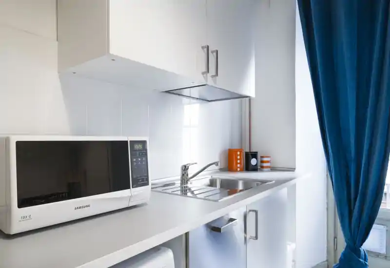 Compact modern kitchenette with a white countertop, built-in sink, microwave and bright natural light from a window with blue curtains.