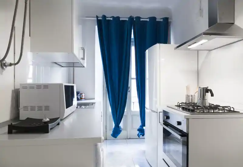 Bright, compact modern kitchen with white cabinets, gas hob, built-in oven and a tall fridge, accentuated by deep blue curtains and natural light.