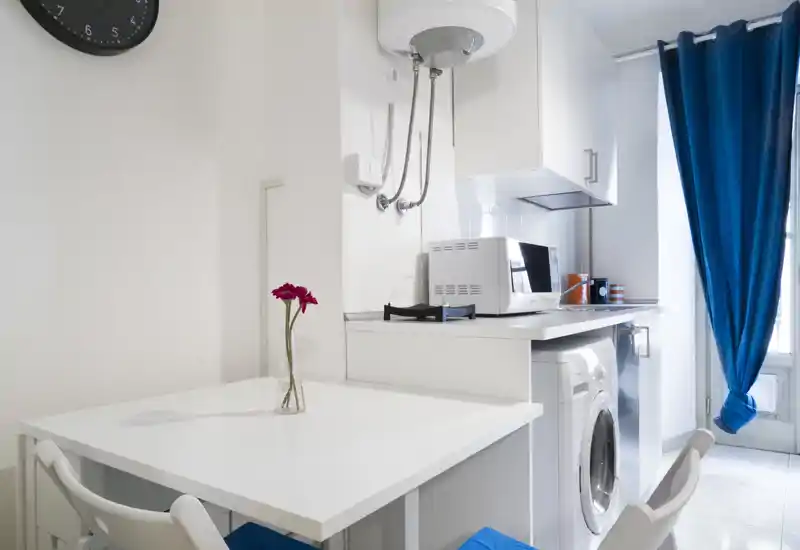 Compact, modern kitchenette with a white dining table, microwave, washing machine and bright natural light from a window. Simple, functional layout ideal for short stays.