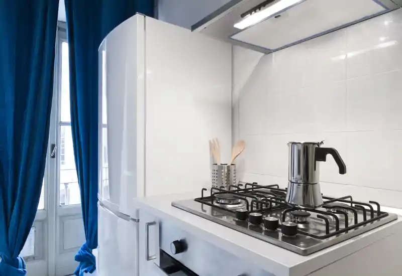 Bright modern kitchenette with a gas hob, stainless stovetop coffee maker, fridge and natural light from a window — compact and well maintained.