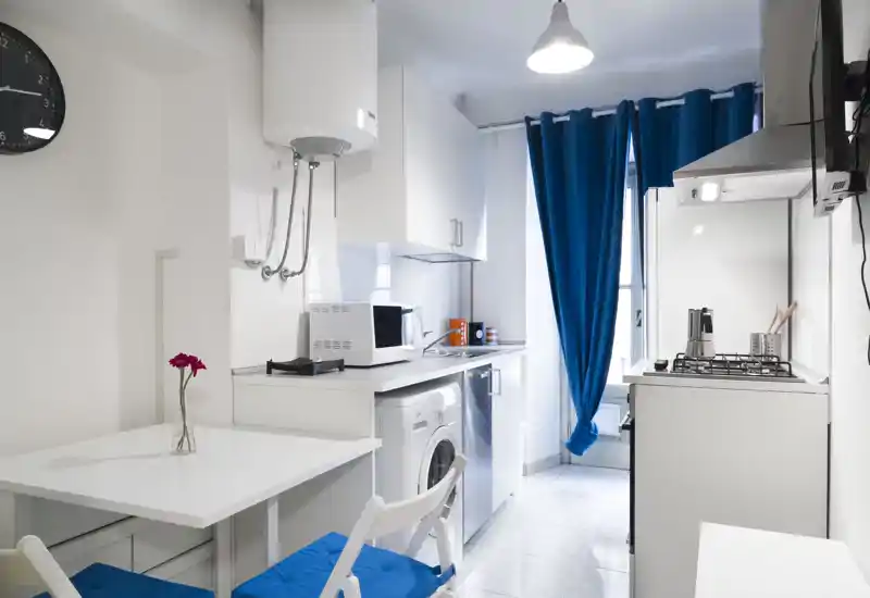 Compact modern kitchen with white cabinets, a gas stove, microwave, washing machine and a bright blue curtain; clean, functional layout ideal for a small apartment.