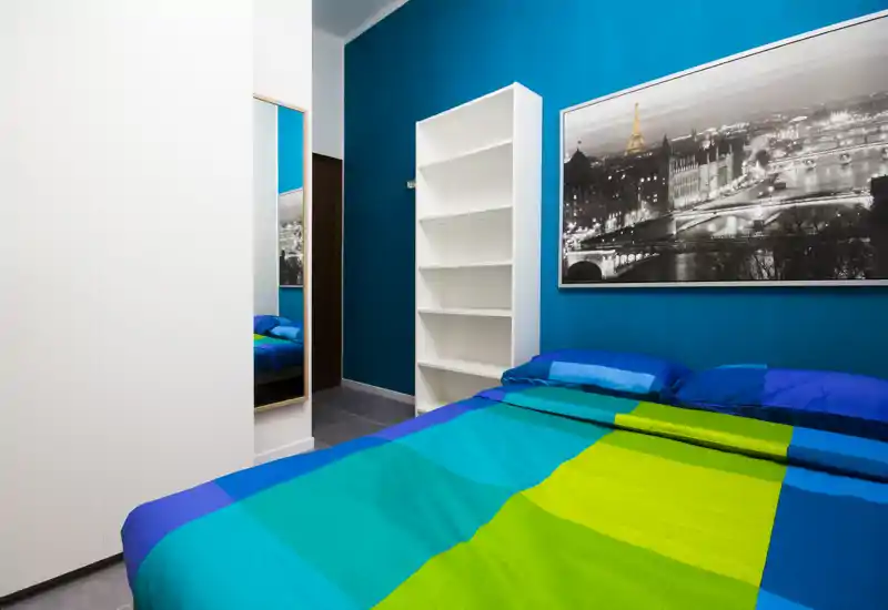Dormitorio moderno y luminoso con ropa de cama colorida, gran fotografía enmarcada y estantería abierta; pared de acento turquesa que aporta un ambiente fresco y acogedor.