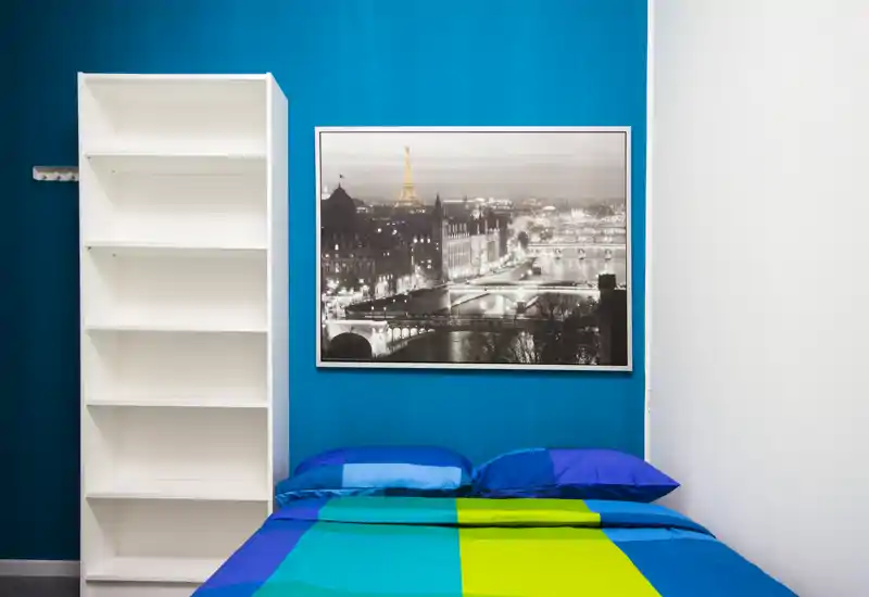 Dormitorio moderno y luminoso con cama doble colorida frente a una pared azul intensa y una gran foto enmarcada de la ciudad; estantería blanca para almacenamiento.