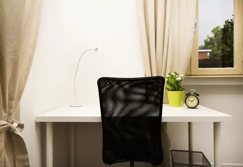 Espacio de trabajo luminoso y minimalista con escritorio blanco, silla de oficina de malla, pequeña planta y reloj despertador junto a una ventana con cortinas beige.