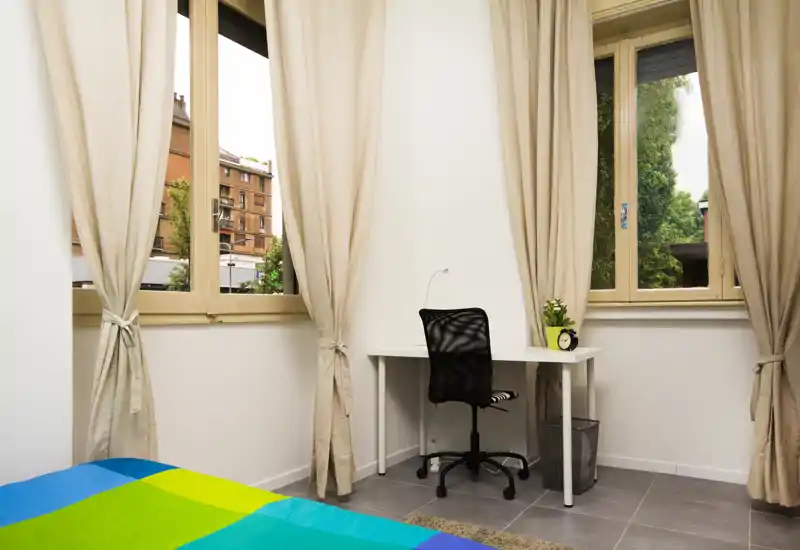 Dormitorio luminoso y ordenado con una cama colorida en primer plano, dos grandes ventanas con cortinas beige y un escritorio compacto con silla — ideal para una estancia cómoda y teletrabajo.