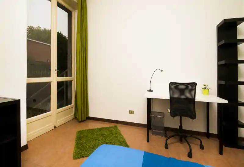 Rincón de dormitorio con cama individual parcialmente visible en primer plano, escritorio y silla junto a una puerta de cristal luminosa y una estantería alta.