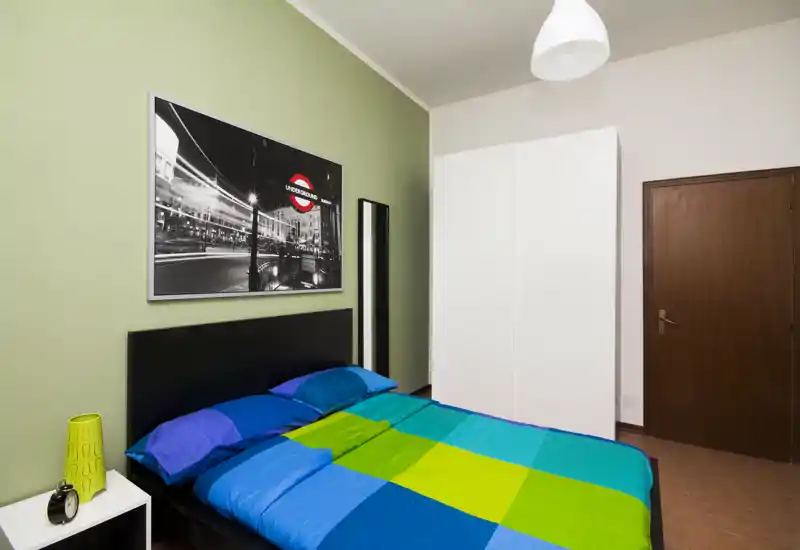 Dormitorio moderno y luminoso con cama doble vestida en tonos azul y verde, cabecero negro, armario y mesita sencilla — ideal para alquileres de corta estancia.