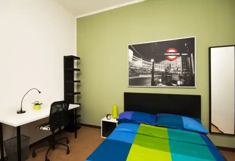 Dormitorio luminoso y decorado con cama doble y ropa de cama colorida, mesita, escritorio y estantería; ideal para estudiantes o una persona.