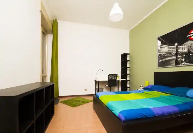 Dormitorio moderno y luminoso con cama doble y colcha colorida, escritorio con silla, estanterías y pequeña alfombra — limpio, bien iluminado y de mobiliario sencillo.
