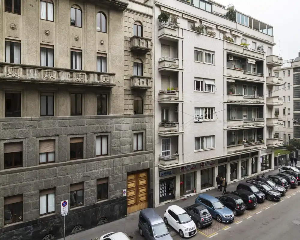 Street-level exterior view of residential buildings with parked cars along the curb and balconies above small ground-floor shops.