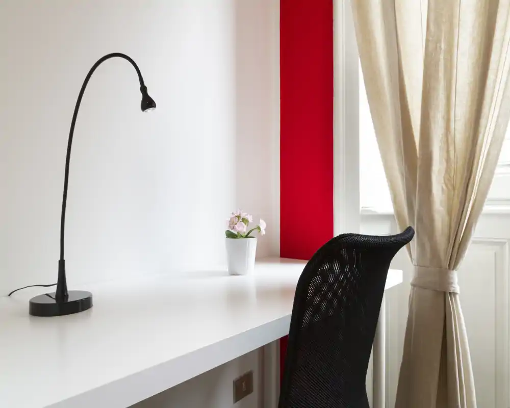 Bright, minimalist workspace with a white desk, black ergonomic chair, task lamp and a small potted plant next to a sunlit window with beige curtains.