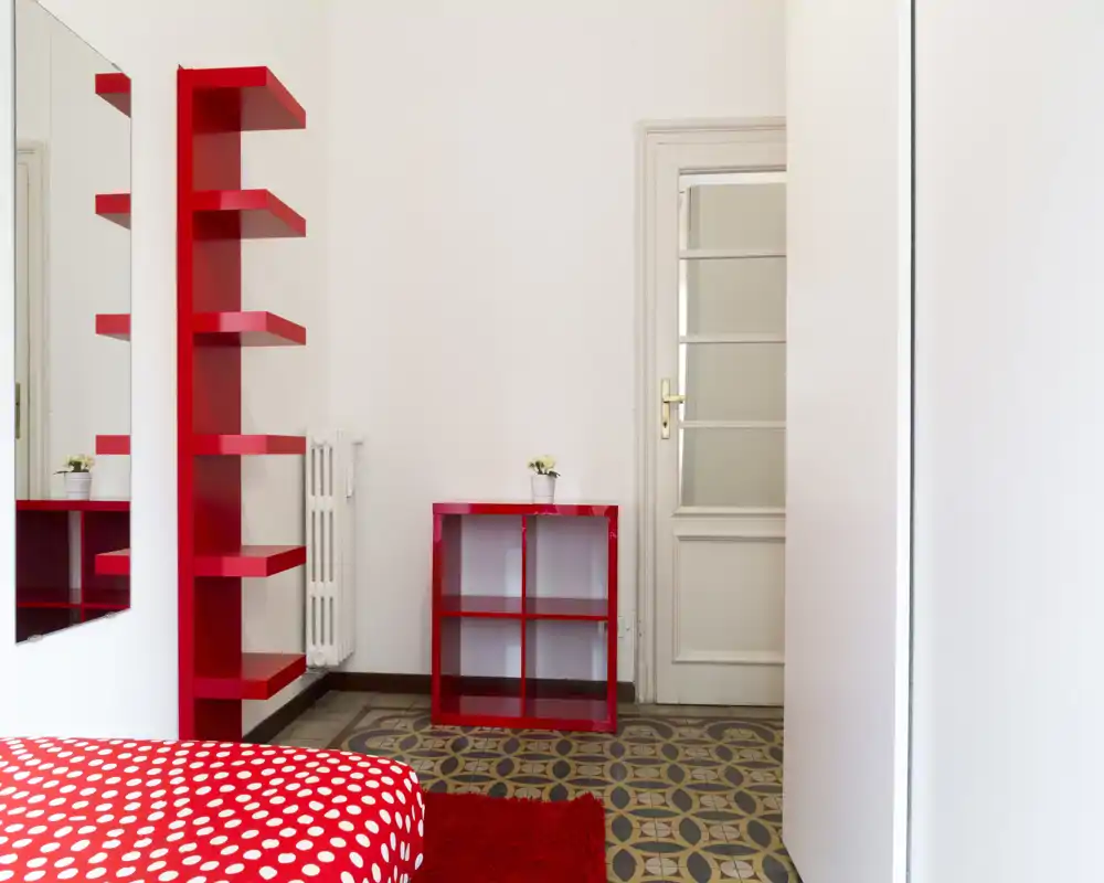 Bright, modern bedroom corner with a red polka-dot bedspread, matching red shelving and a small cubed storage unit on patterned tile flooring.