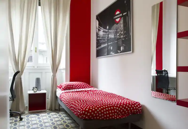 Bright, well-staged single bedroom with a red polka-dot bed, natural light from a large window, patterned floor tiles and a small desk area — clean and inviting for short stays.