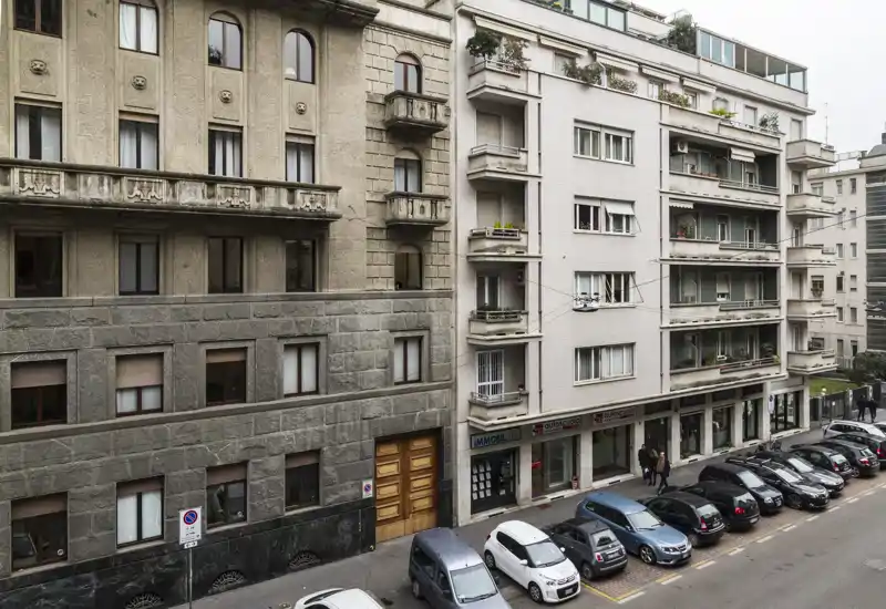 Street-level exterior view of residential buildings with parked cars along the curb and balconies above small ground-floor shops.