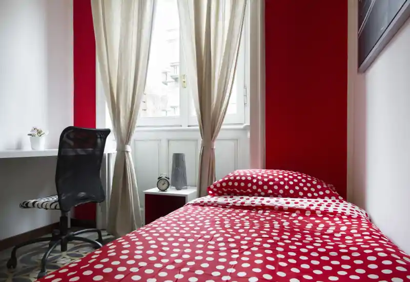 Bright, compact bedroom with a single bed dressed in red polka-dot bedding, a small bedside table and a desk by the window — ideal for a solo traveler or student.