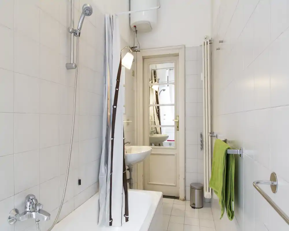 Bright, clean bathroom with a bathtub and showerhead, small sink, towel rail and mirrored door; white tiles create a fresh, minimalist look.