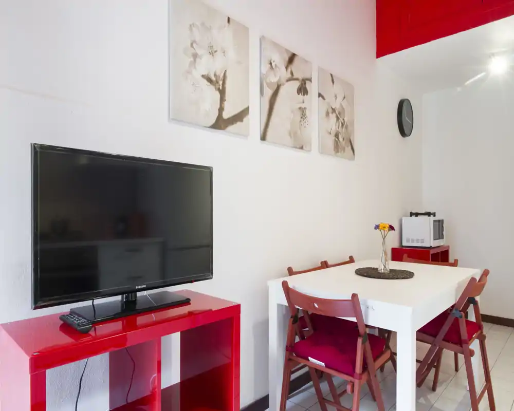Bright, modern dining area with a white table, four red-cushioned chairs and decorative wall art; a TV on a red console and a vase with flowers complete the space.