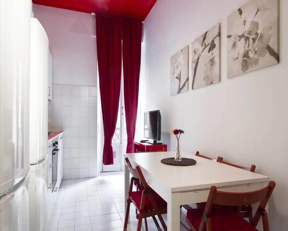Bright, compact dining area with a white table, red folding chairs and coordinated red accents including curtains and ceiling; natural light comes from a glass door, and a small kitchen is visible beyond.