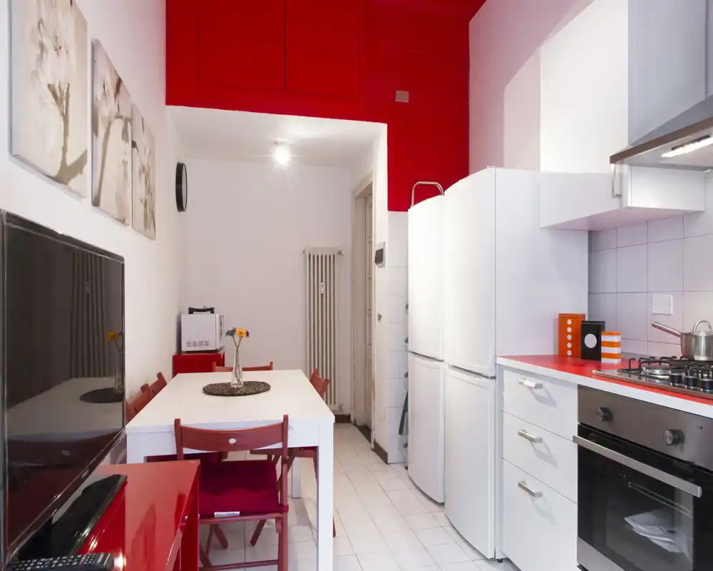 Bright compact kitchen with integrated dining area. White cabinets and appliances contrast with bold red accents and a simple table set for four, creating a modern, welcoming space.