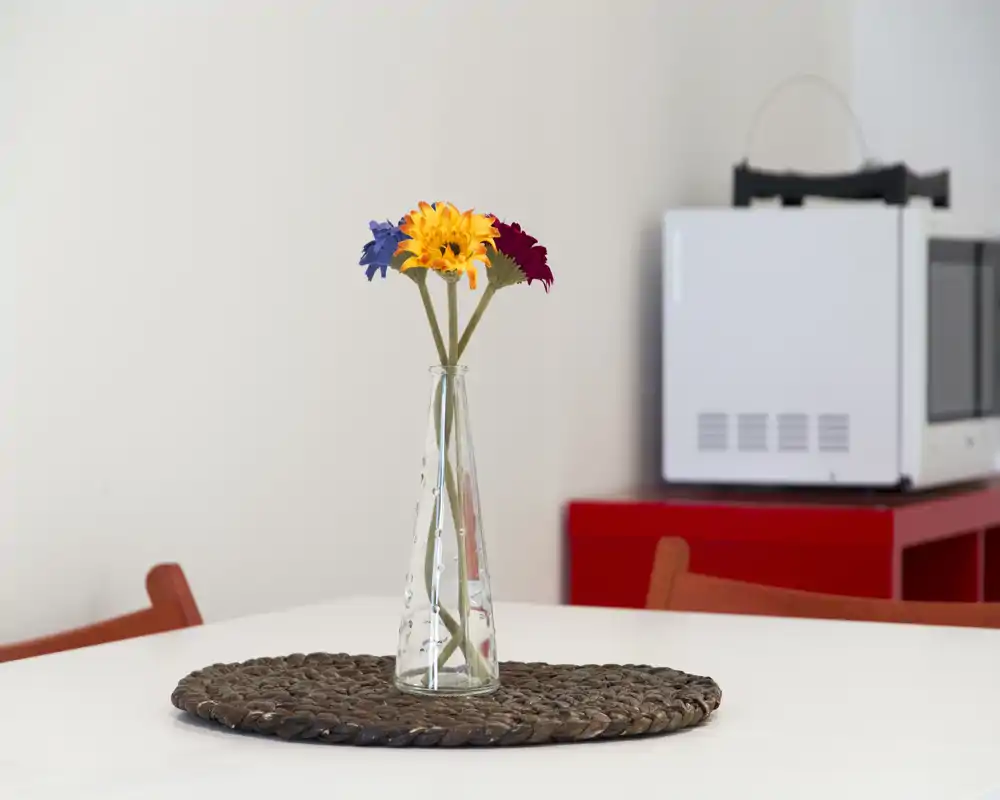 A minimal dining table scene with a glass vase holding three colorful flowers as a simple centerpiece on a white table.